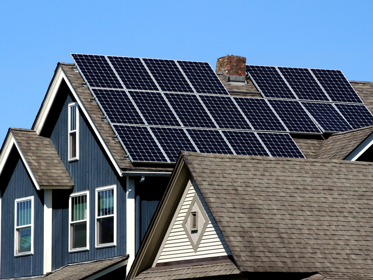 Residential house with multiple solar panels mounted across the sloped roof, with clear blue sky visible in the background.